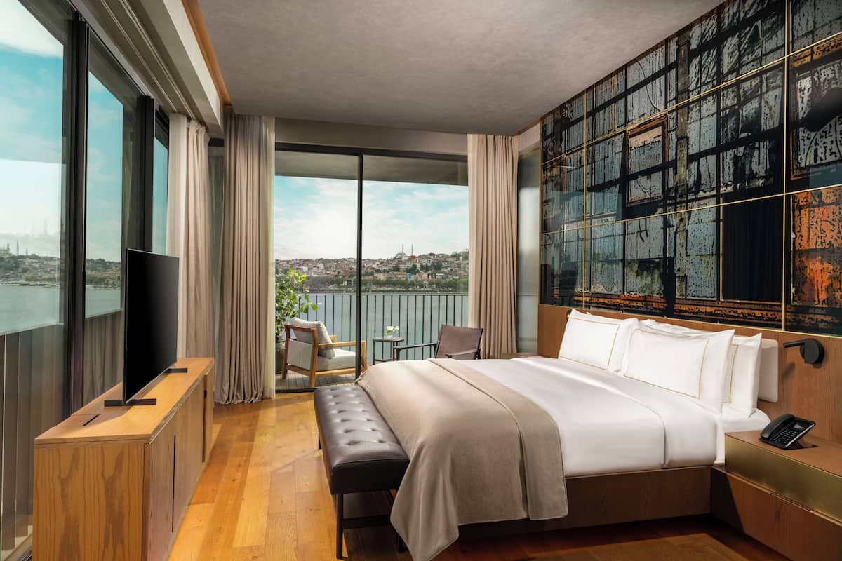 Habitación de hotel moderna con una cama grande, un televisor, un banco y vistas a un paisaje urbano a través de grandes ventanales.