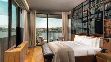 Habitación de hotel moderna con una cama grande, un televisor, un banco y vistas a un paisaje urbano a través de grandes ventanales.