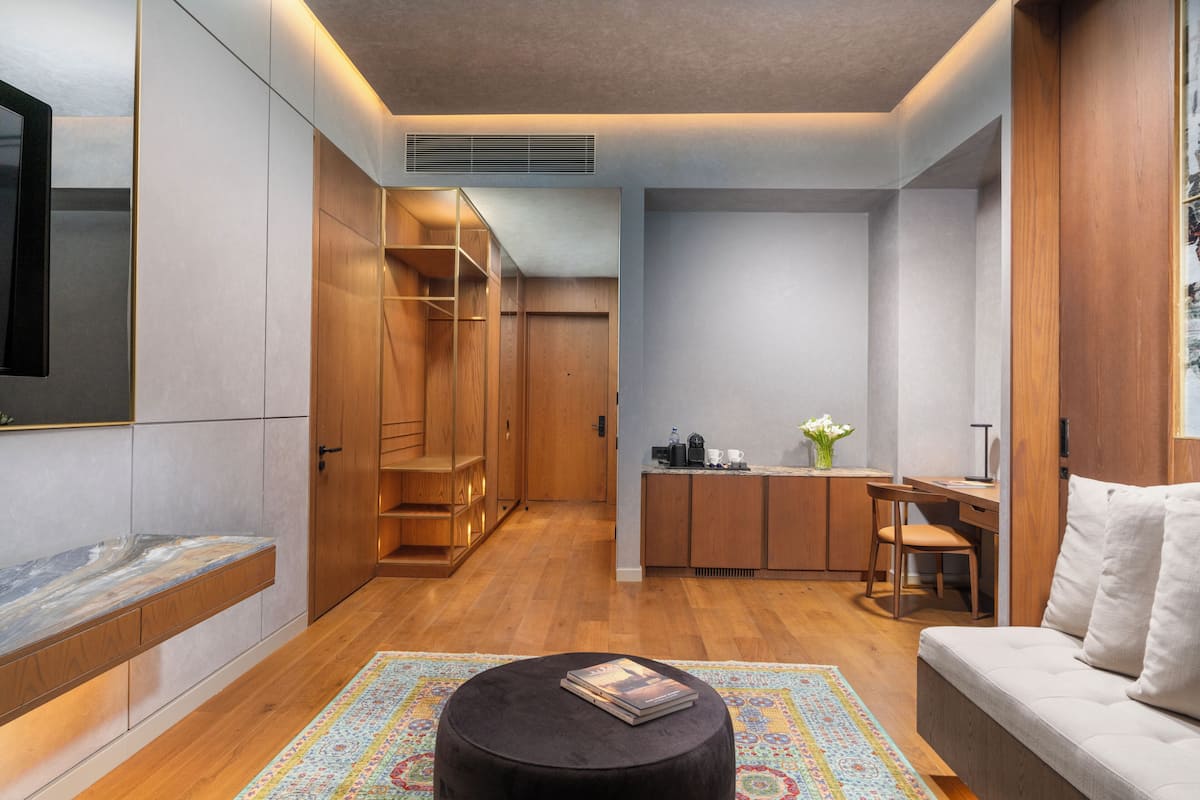 Un interior moderno con sofá, reposapiés y una mesita con un libro. Se observa una zona de cocina con fregadero, estufa y armarios. También se ve un armario de madera y un televisor.