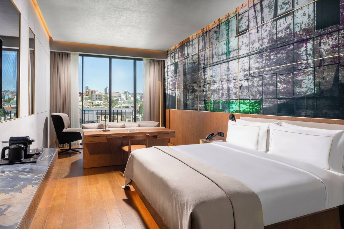 Una habitación de hotel moderna con una cama grande, un escritorio con una silla, vistas de la ciudad a través de amplias ventanas y una pared con una textura original.