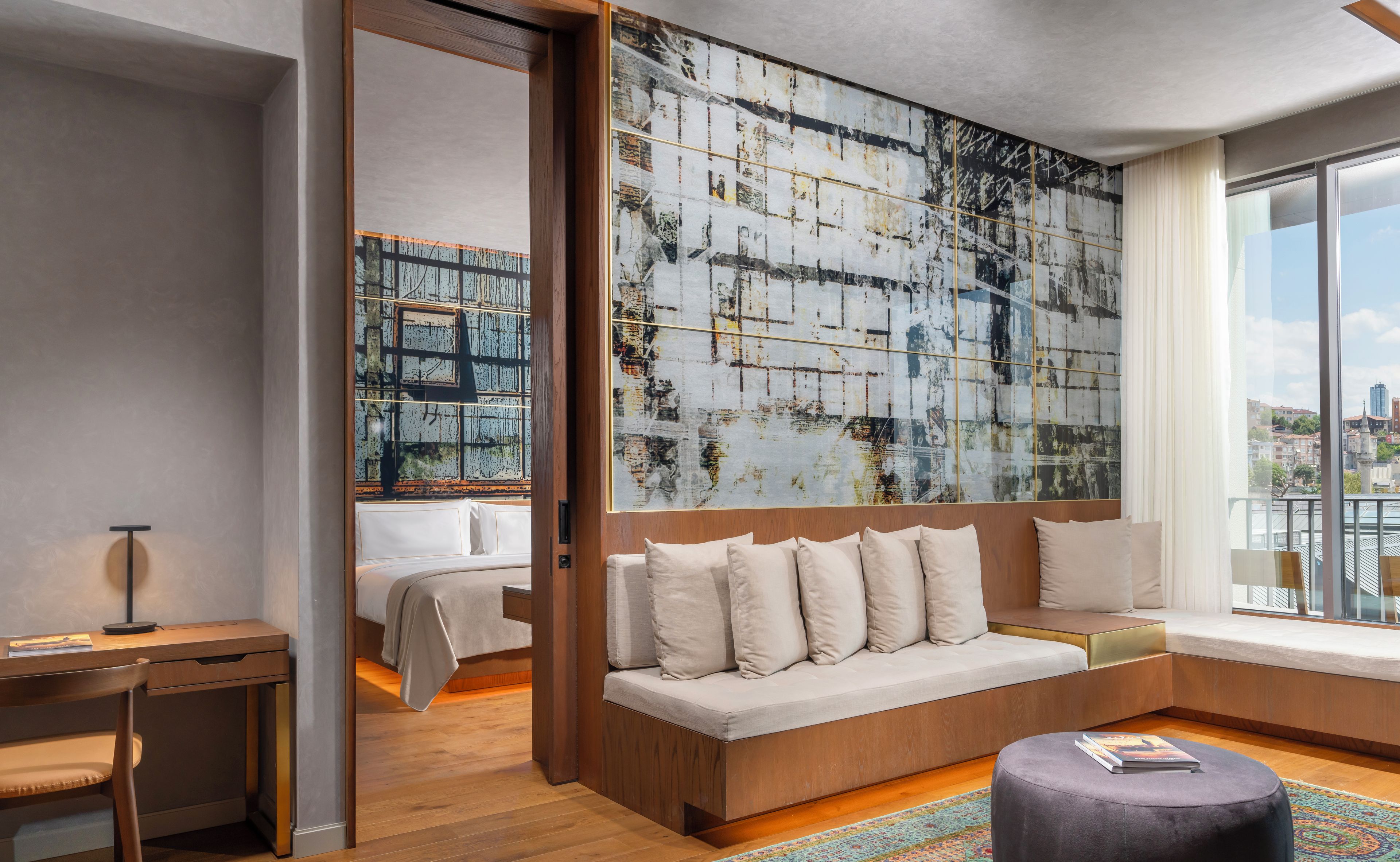 Habitación de hotel moderna con un cuadro abstracto grande, un sofá con varios cojines, un escritorio y vistas de la ciudad a través de amplias ventanas.