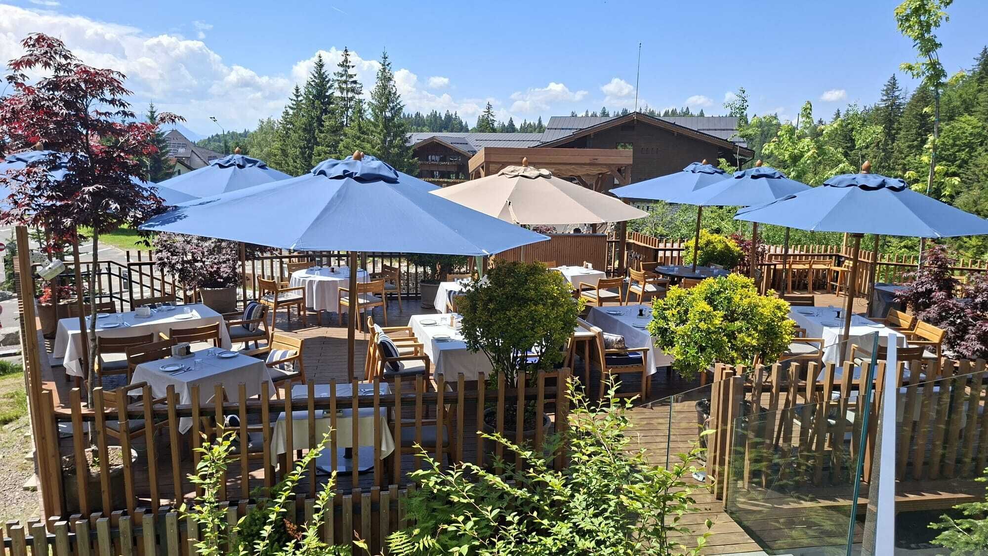 Un área de comedor al aire libre con mesas y sillas, cubierta por sombrillas y rodeada de vallas de madera y follaje.