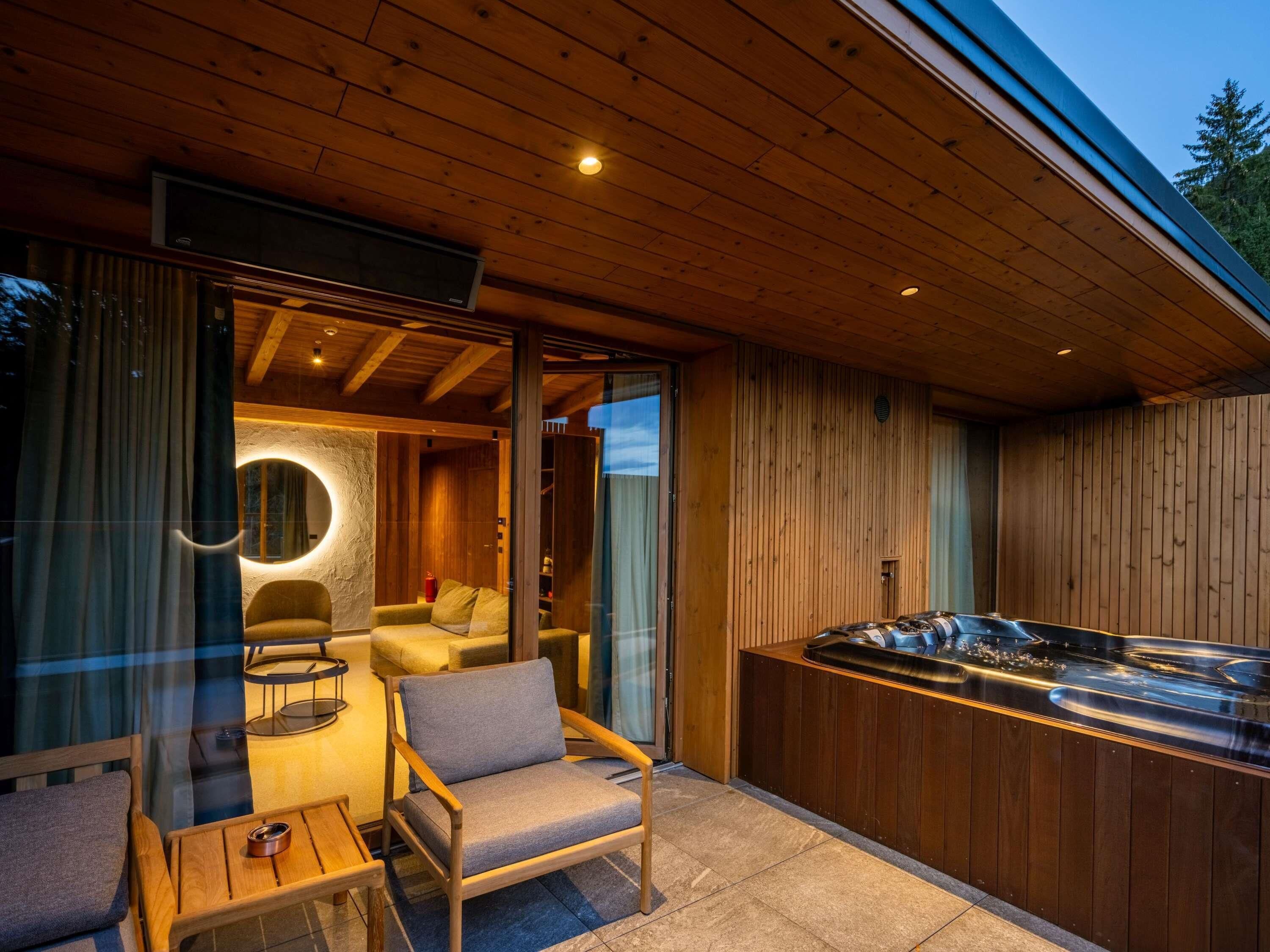 A modern outdoor seating area with a hot tub, wooden furniture, and a glass wall.