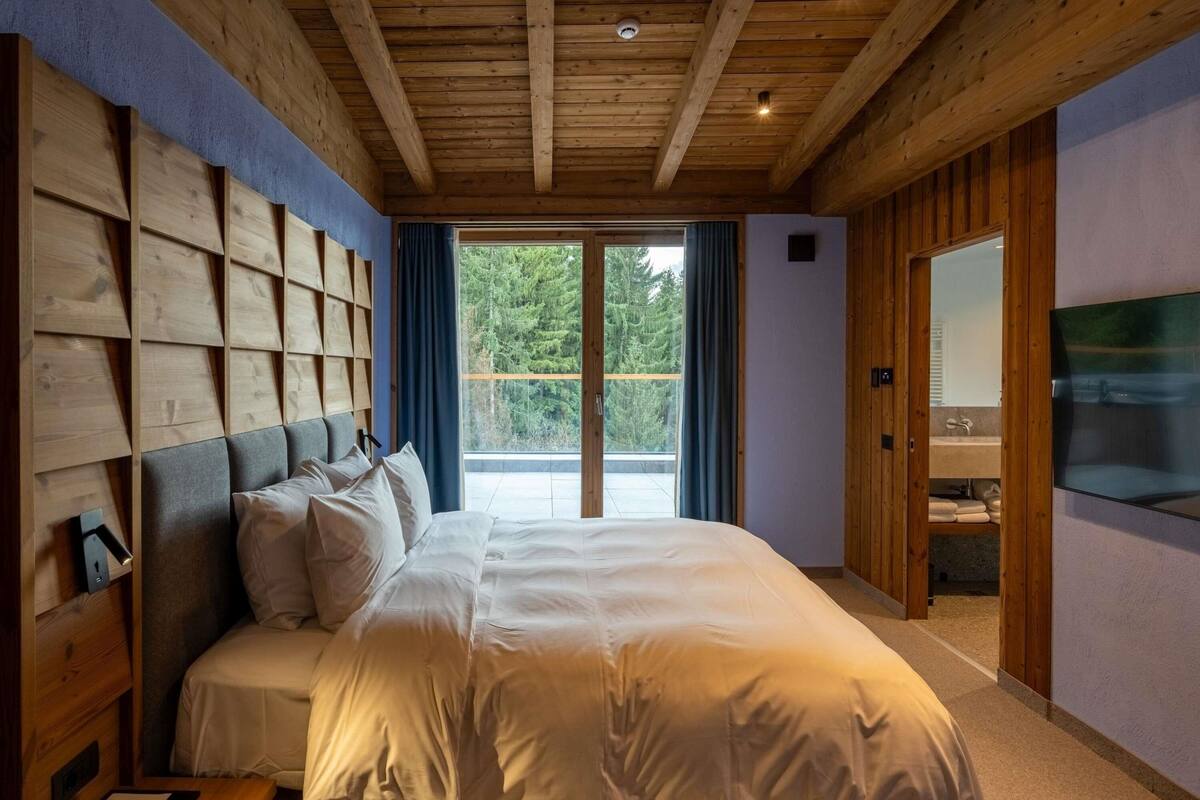 Ein Schlafzimmer mit einem großen Bett, einem Holzkopfteil, einem Fernseher und Blick auf Bäume durch ein Fenster.