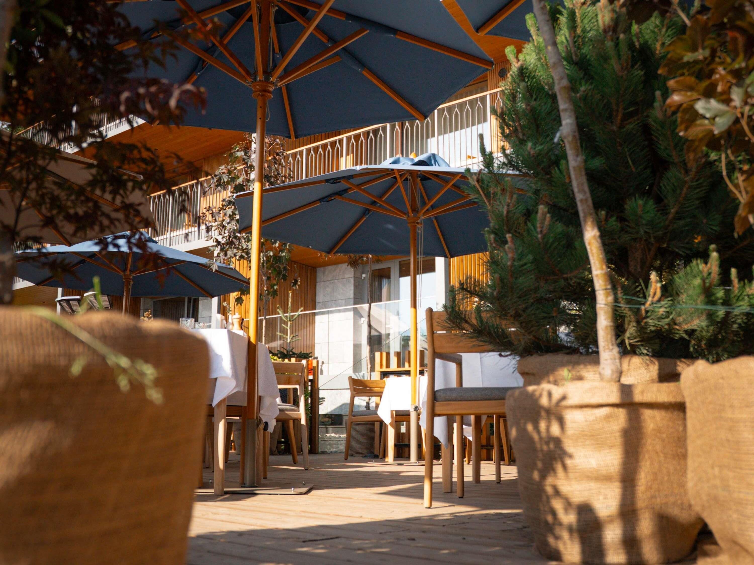 An outdoor dining area with wooden tables and chairs, umbrellas, and potted plants.