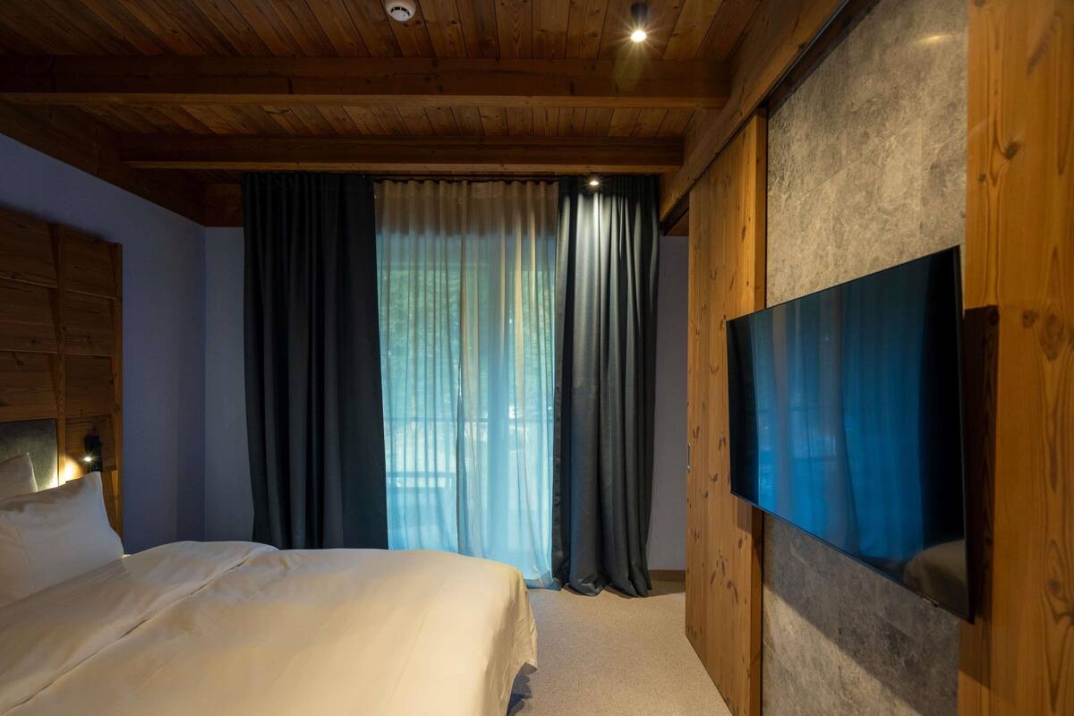 Ein Schlafzimmer mit einem Bett, einem an einer Holzwand montierten Fernseher und einem Fenster mit Vorhängen.