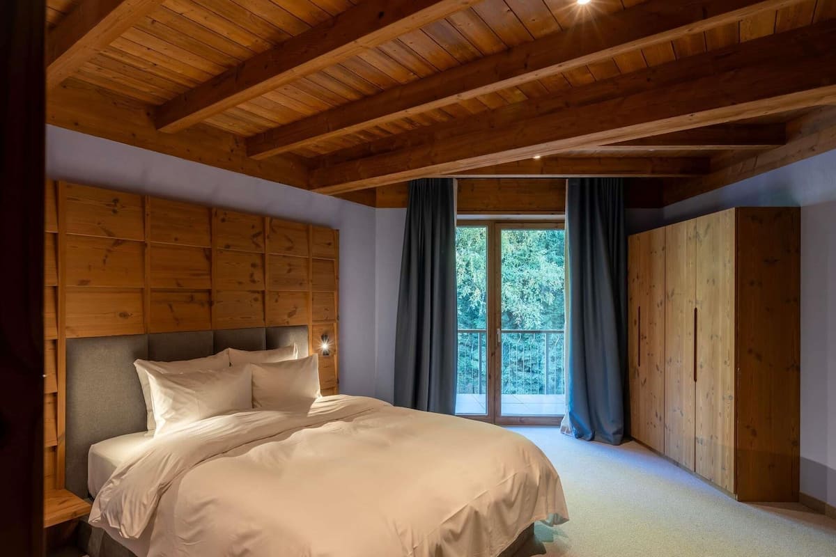 Un dormitorio con techo de madera, una cama grande con ropa de cama blanca, cabecera de madera y vistas a árboles a través de un ventanal.