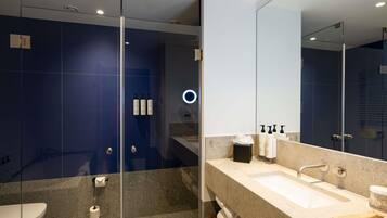 Un baño moderno con cabina de ducha de vidrio, un espejo grande y una encimera color beige con lavabo y artículos de aseo.