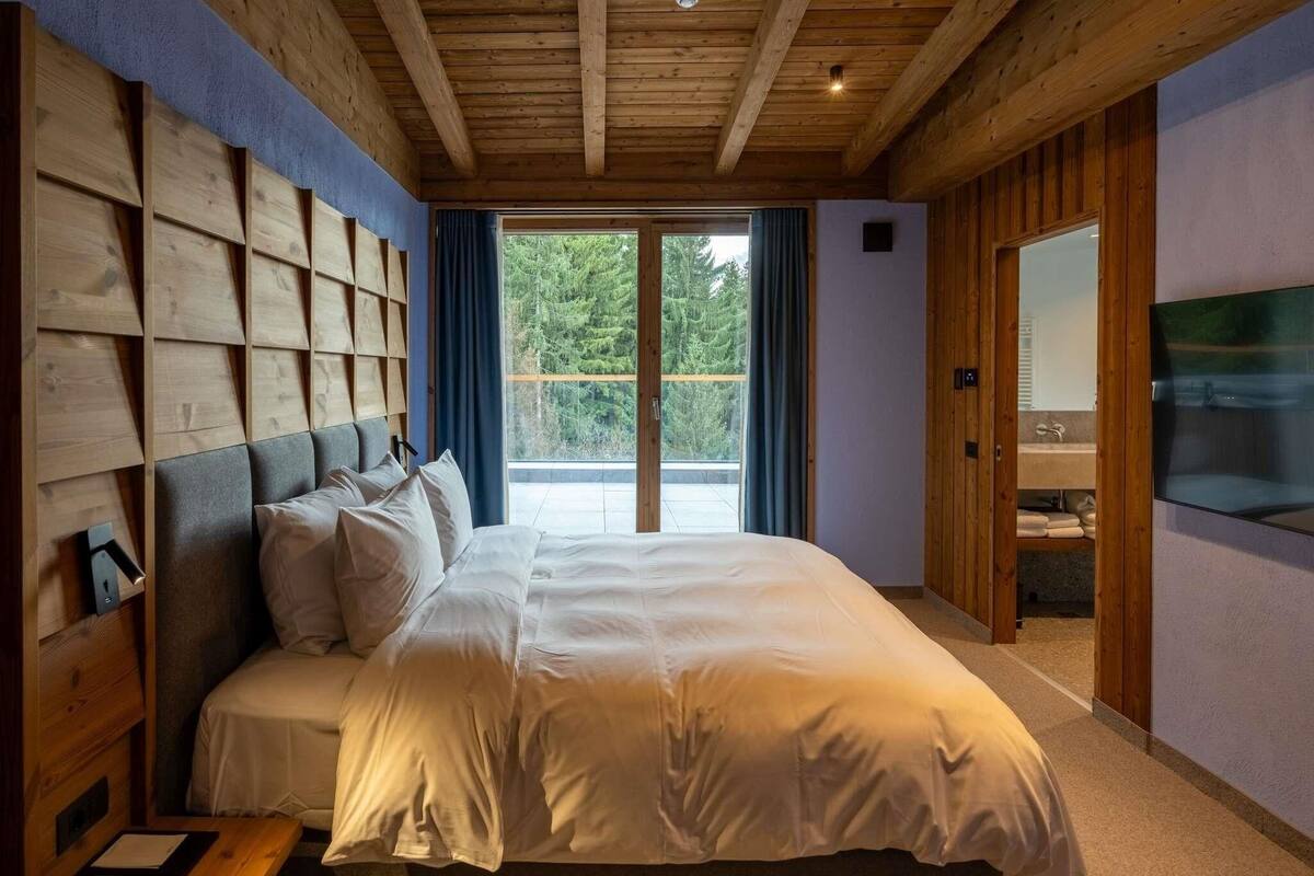 Une chambre avec un grand lit, une tête de lit en bois, une télévision et une vue sur les arbres par la fenêtre.