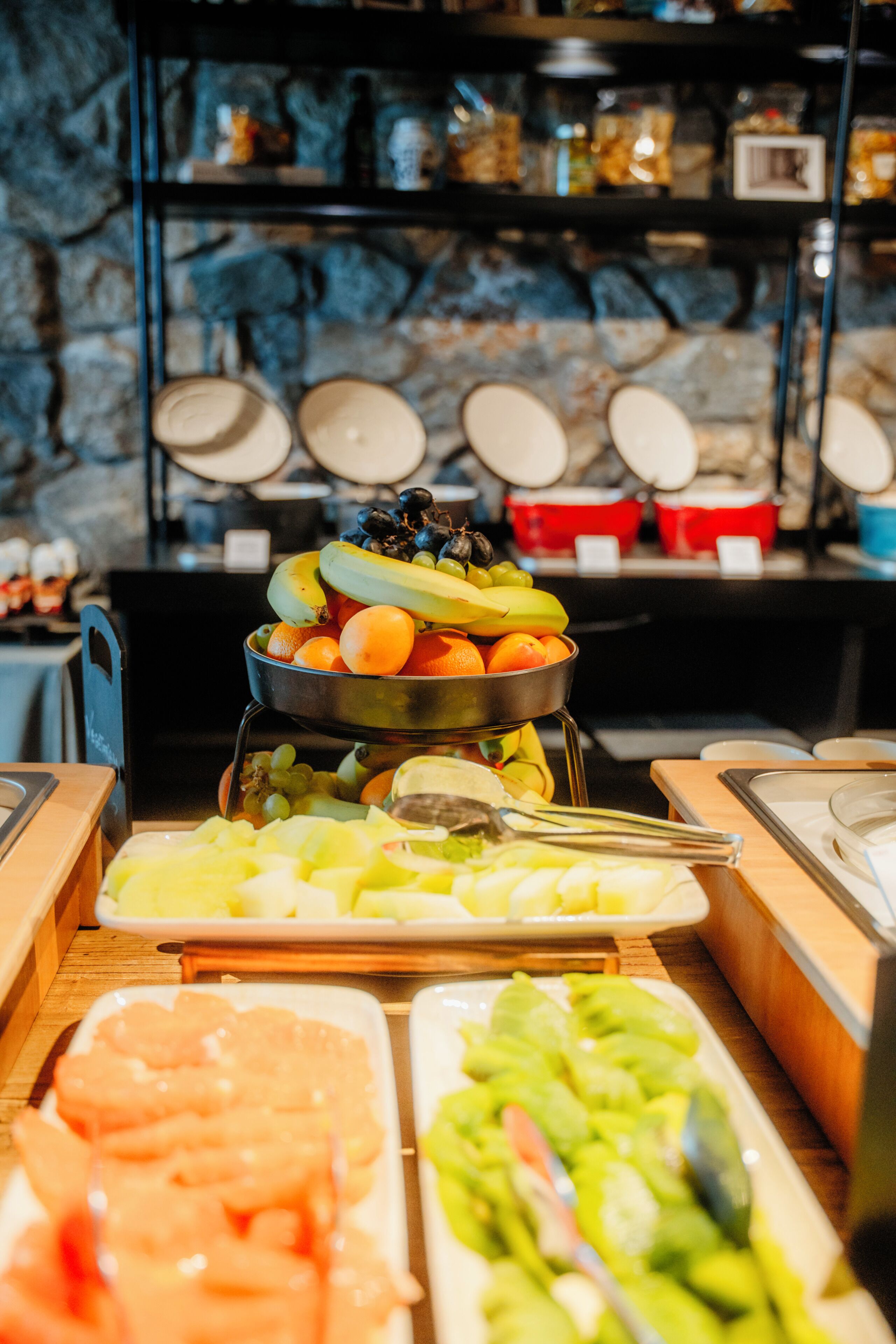A buffet with various fruits and vegetables, a stone wall background, and a metal rack with plates.