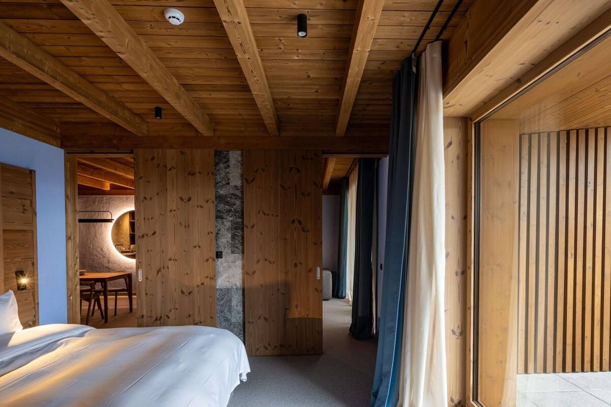 Ein Holzinnenraum mit einem Bett, einem Schreibtisch und einem durch eine Glastür einsehbarer Badezimmer.