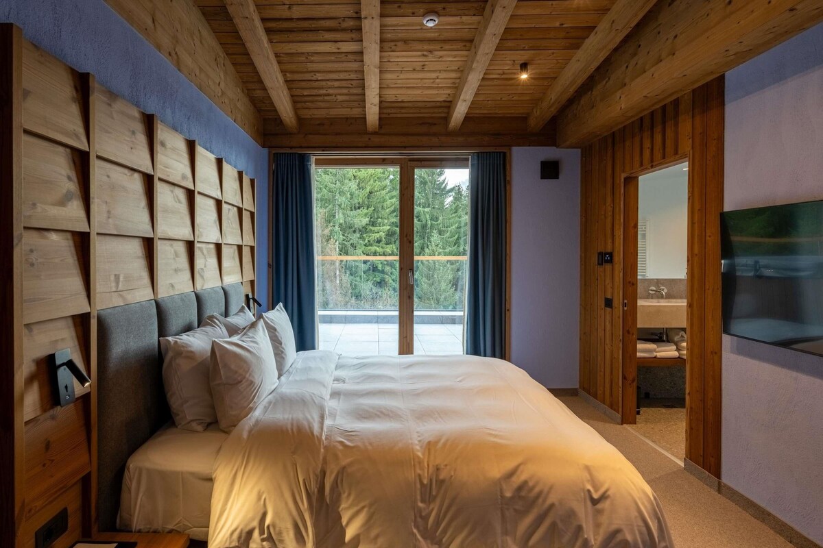 Une chambre avec un grand lit, une tête de lit en bois, une télévision et une vue sur les arbres par la fenêtre.