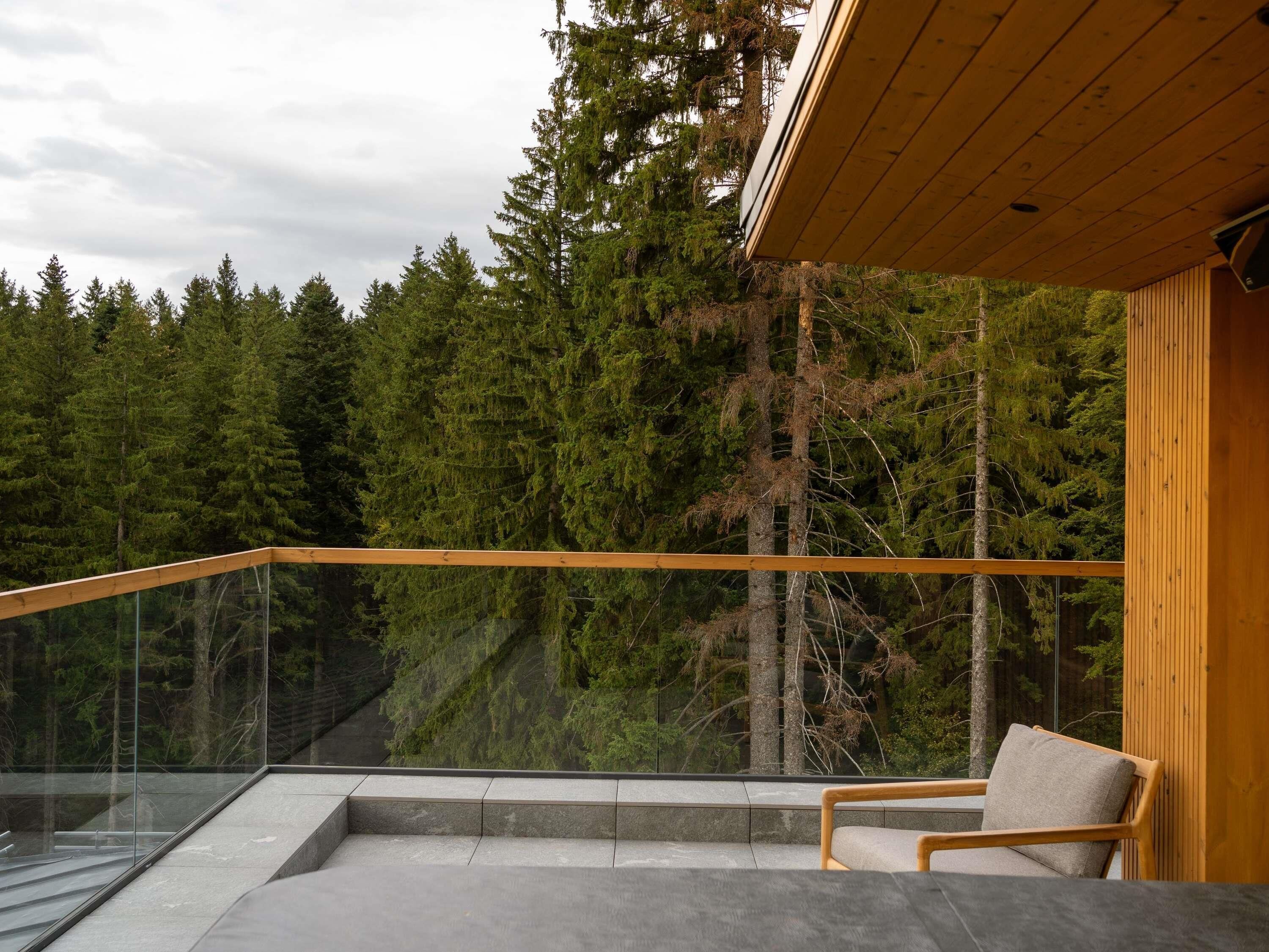 A balcony with a glass railing, a wooden chair, and a view of a forest.