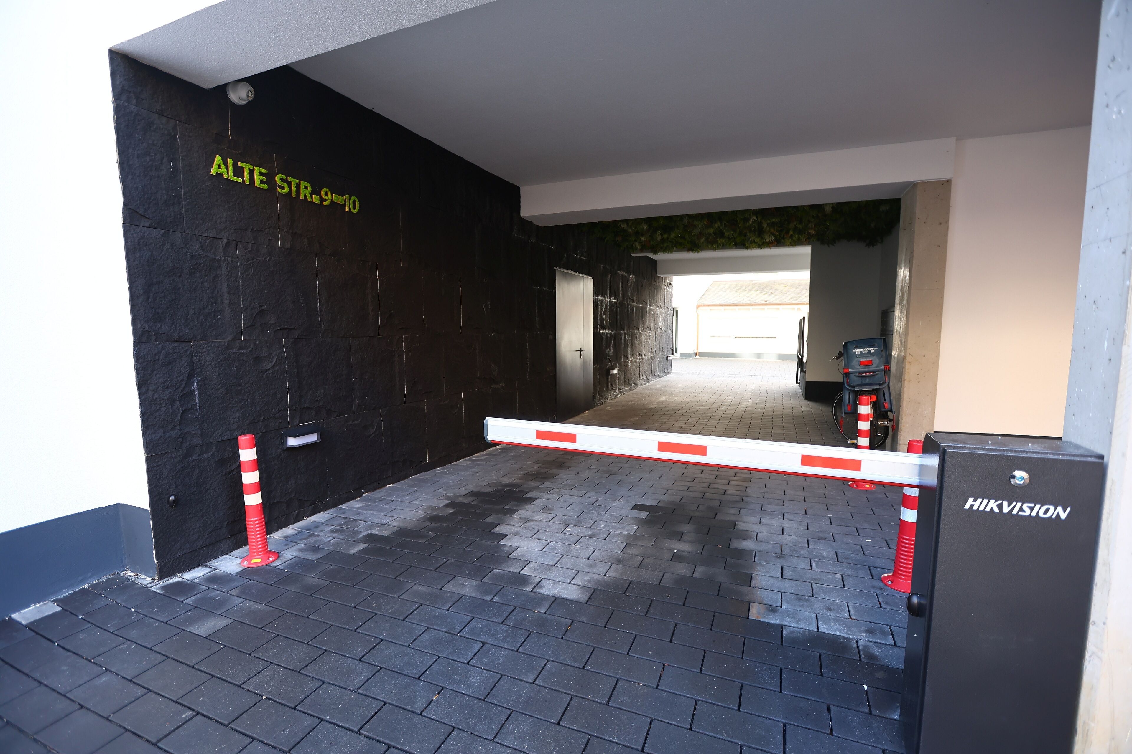 A gated entrance with a black wall, a sign reading "ALTE STR.9-10", and a HIKVISION security box.