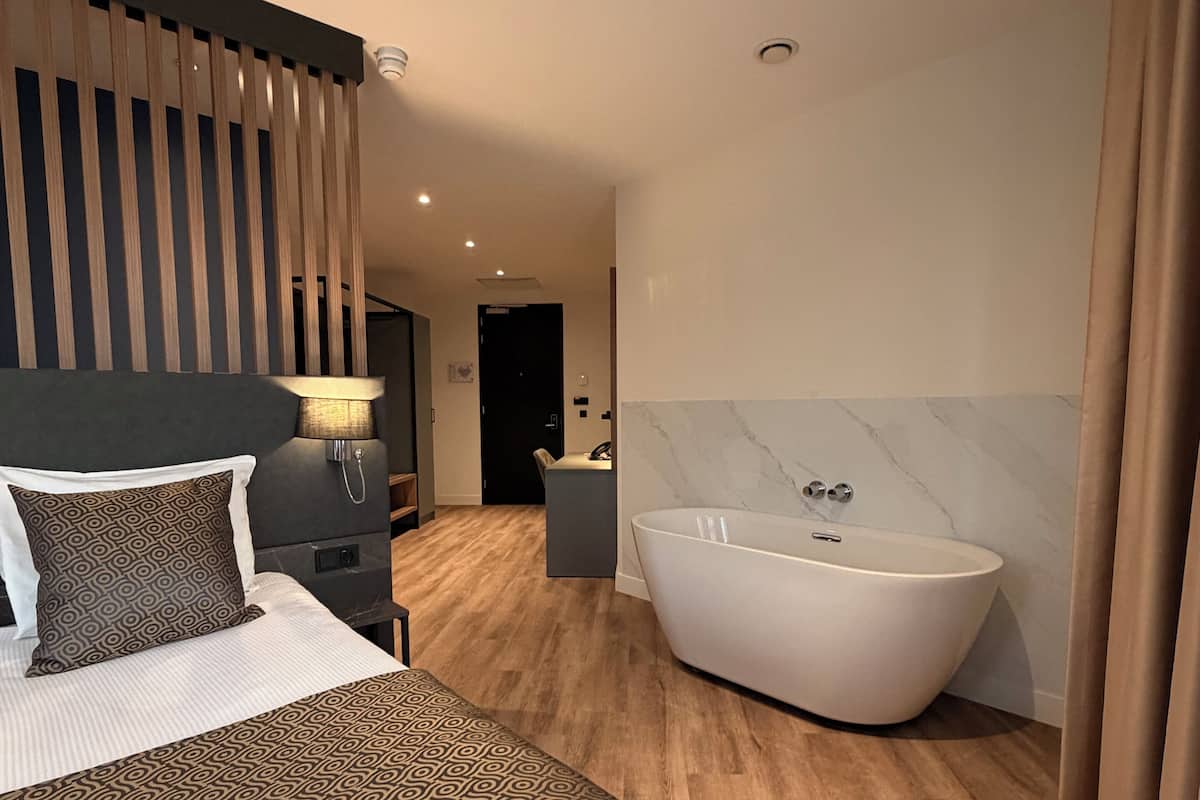 A modern hotel room with a large bed, a freestanding bathtub, and a wooden floor.