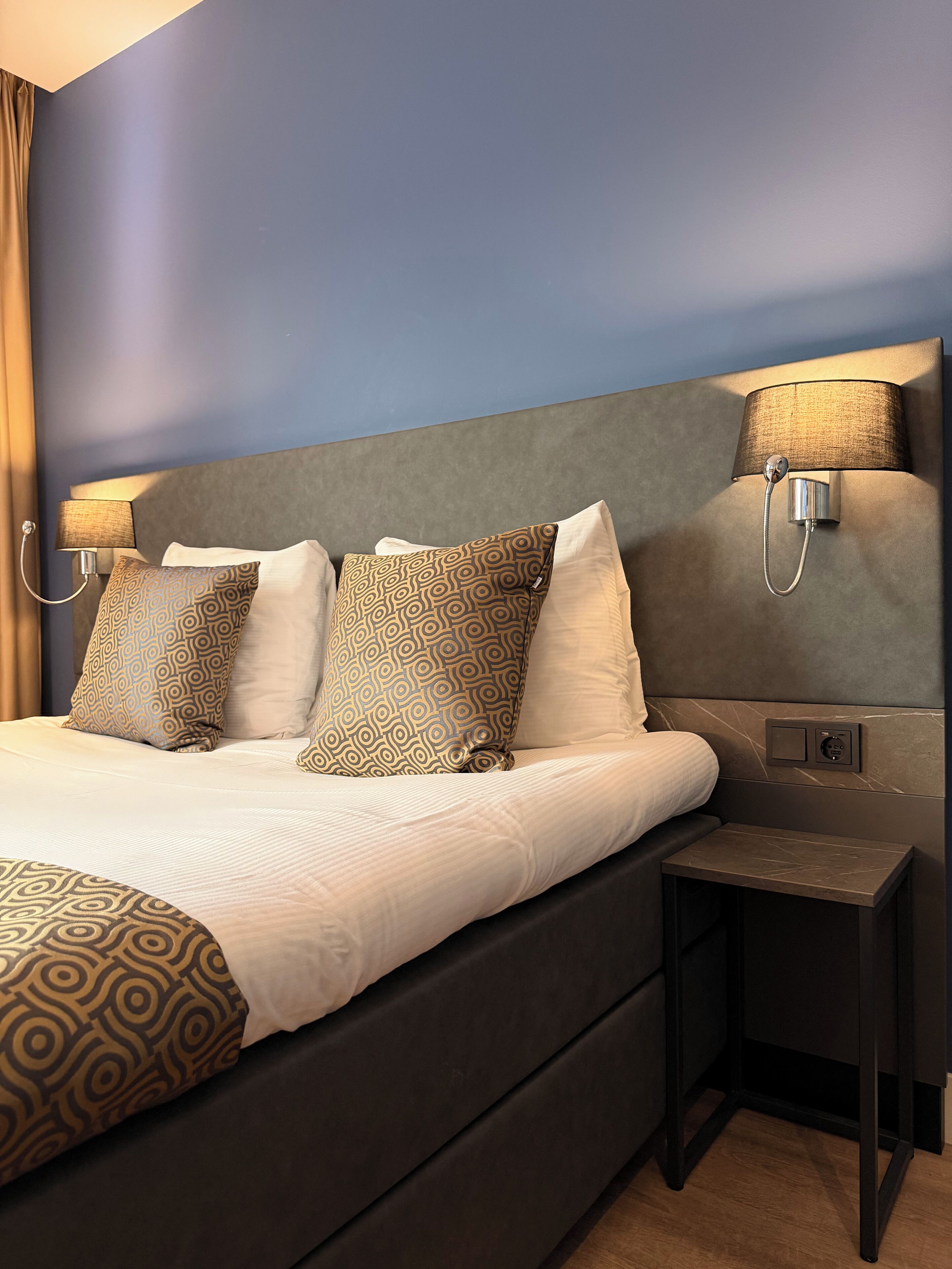 Habitación de hotel con una cama grande, dos lámparas adosadas a la pared, una mesita de noche y un enchufe.