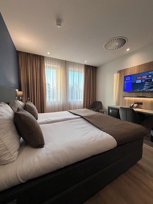 A hotel room with a large bed, a desk, a chair, a TV, and a window with curtains. - OZO Hotels Antares Amsterdam Schiphol Airport (Hoofddorp)