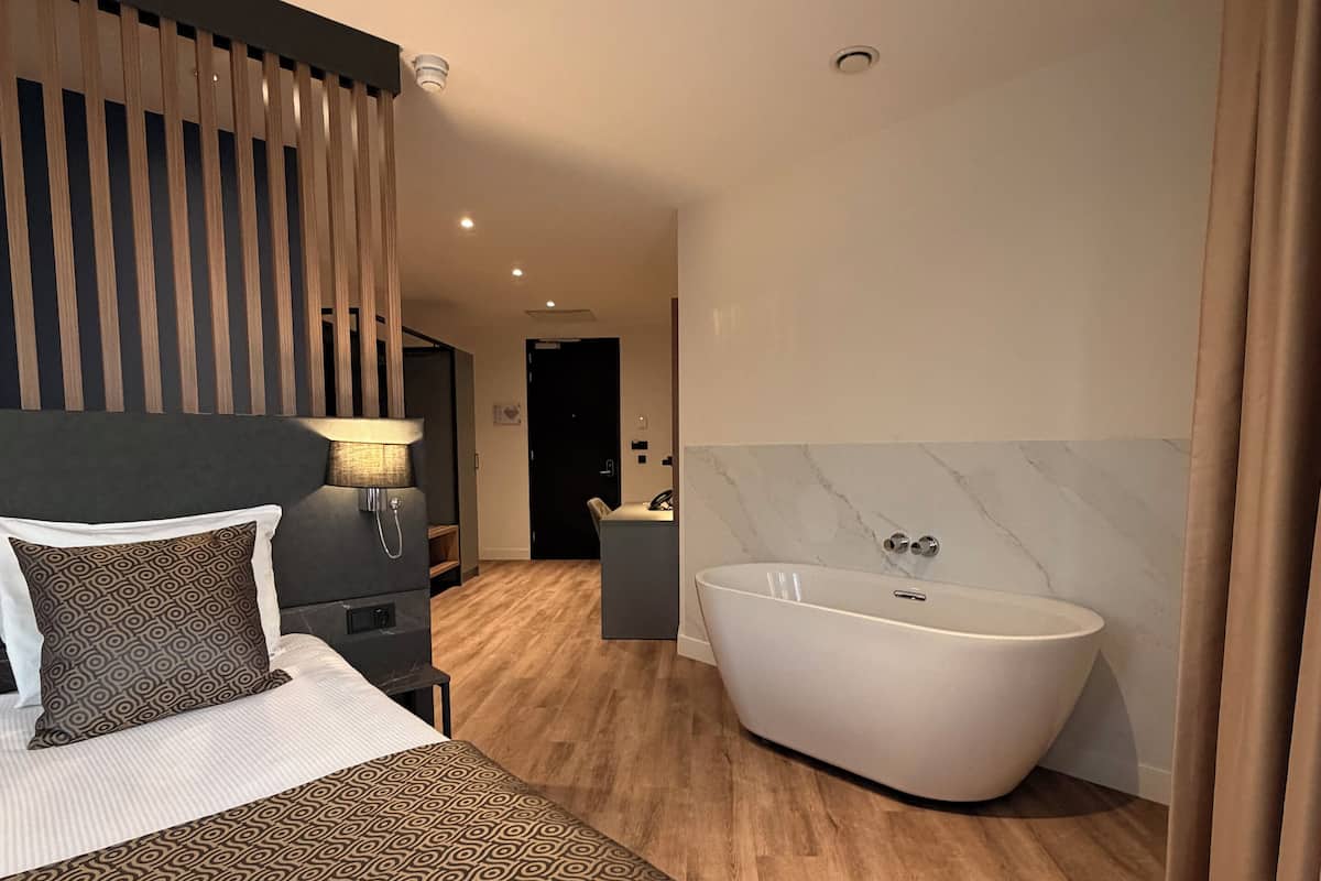 A modern hotel room with a large bed, a freestanding bathtub, and a wooden floor.
