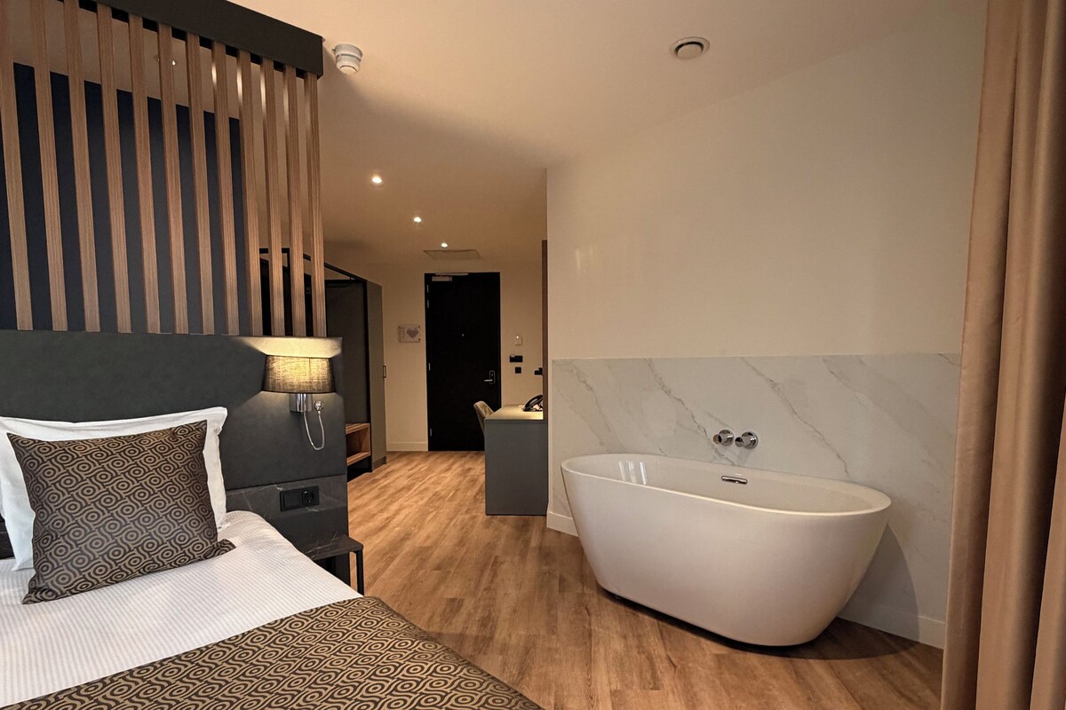 A modern hotel room with a large bed, a freestanding bathtub, and a wooden floor.