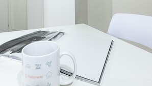 A white table with a mug and a magazine, a white chair, and a door in a room with white walls and a ceiling with beams.