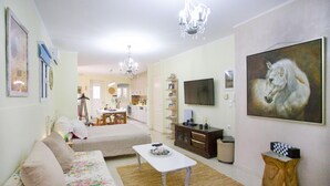 A living room with a sofa, a coffee table, a chandelier, a painting, and a TV on the wall.