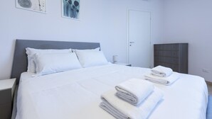 Ein ordentlich bezogenes Bett mit weißen Bettwäsche und Handtüchern, ein grauer Kopfteil, ein weißer Nachttisch, eine graue Kommode und zwei abstrakte Wandbilder.