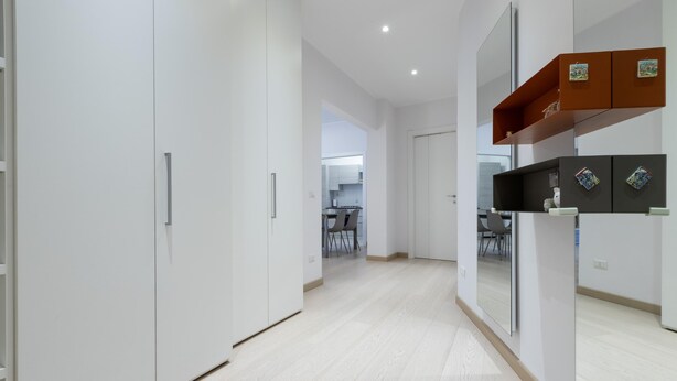 Ein moderner, minimalistischer Flur mit weißen Wänden und Holzboden. Links befinden sich weiße Schränke mit silbernen Griffen, rechts ein Holzregal mit Dekorationsobjekten, und im Hintergrund ist ein Blick in einen Küchenbereich erkennbar.