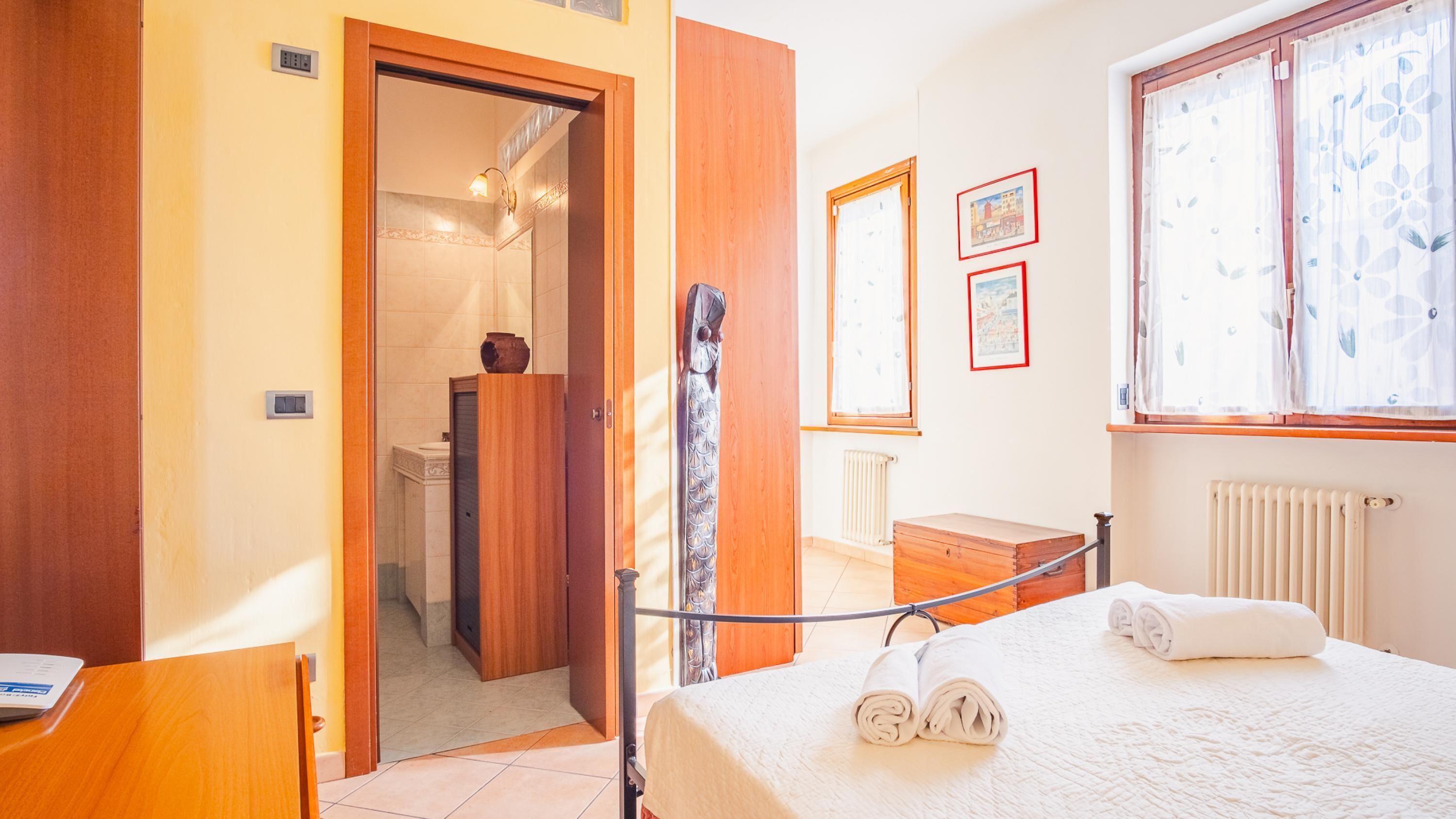 A hotel room with a bed, a wooden wardrobe, a small bedside table, a radiator, and a window with curtains.
