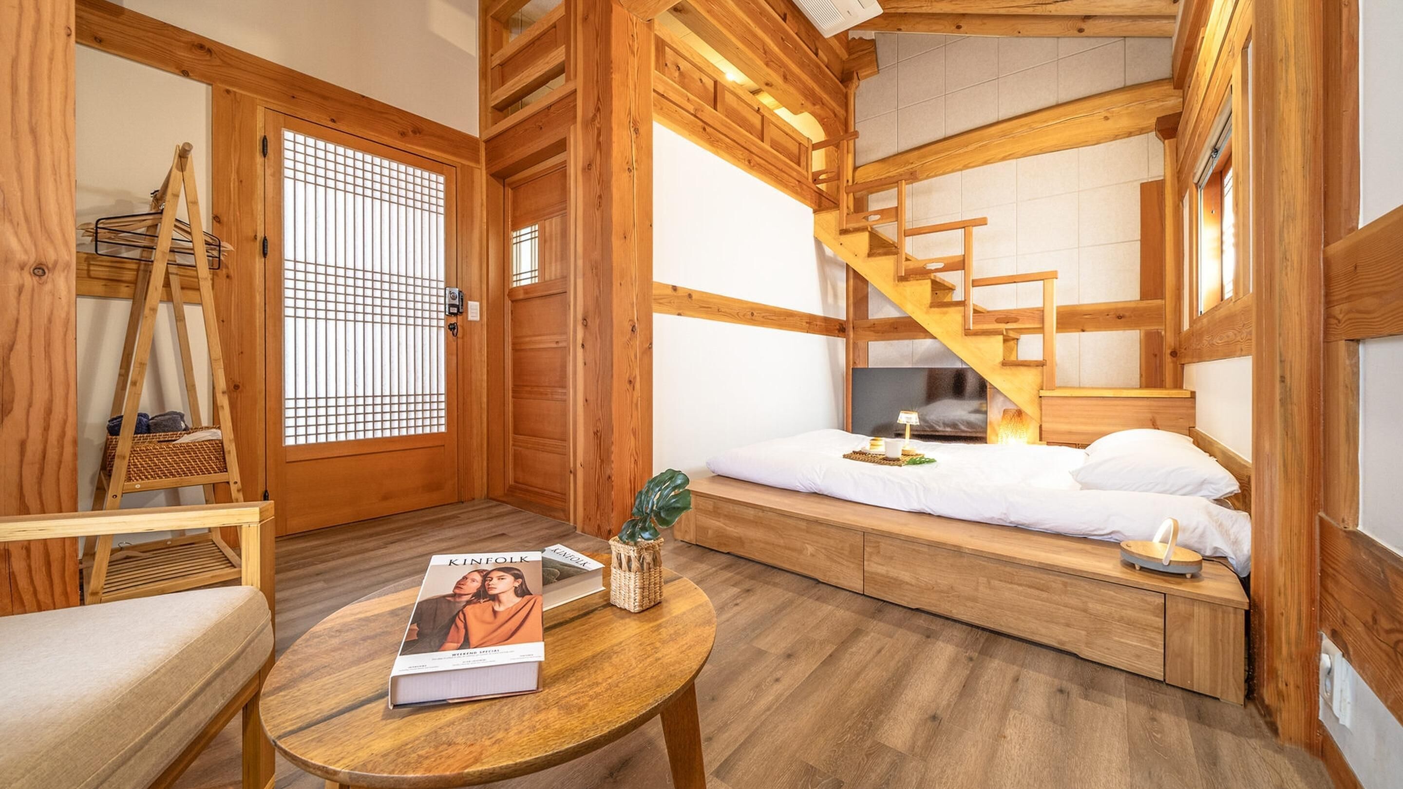 A cozy wooden room with a bed, a small table with a book, a ladder, and a window with a grid pattern.