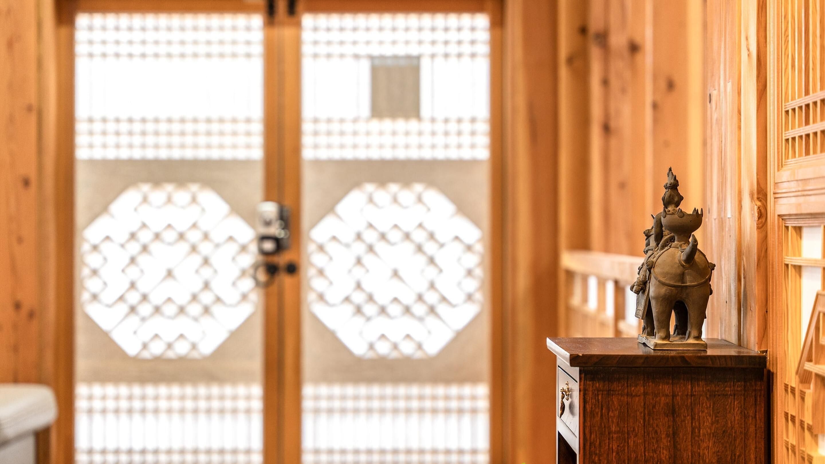A traditional Japanese room with a sliding door featuring a geometric pattern, a wooden dresser with a small statue, and a wooden wall panel.