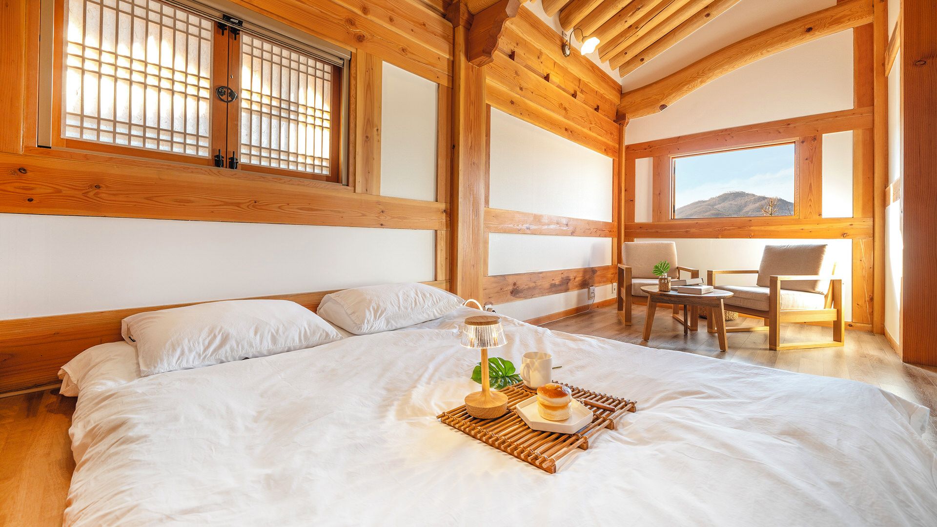 A room with a large bed, wooden beams, a view of the outdoors, and a small table with a lamp.