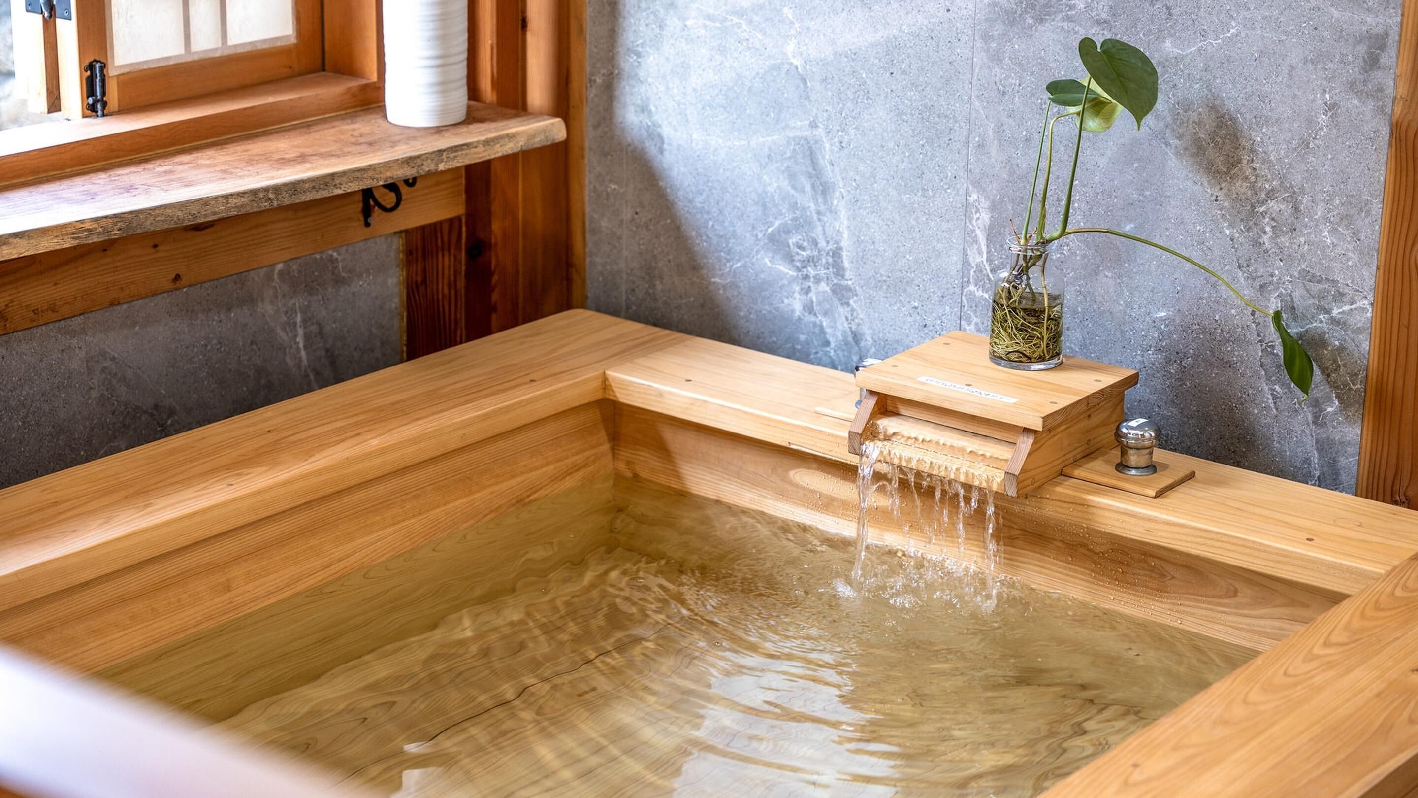 A wooden hot tub with a small waterfall feature, a potted plant, and a wooden shelf with a white container.