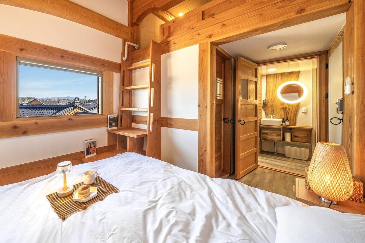 Ein Schlafzimmer mit Holzboden, einem Bett, einem Nachttisch mit Lampe, einem Fenster mit Aussicht und einem Badezimmer mit Spiegel und Waschbecken.