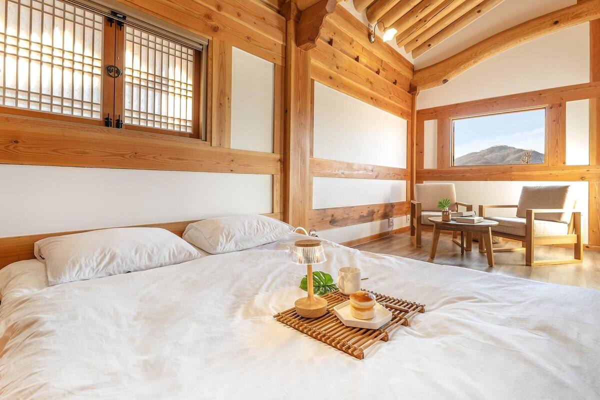 Ein Schlafzimmer mit einem großen Bett, Holzbalken, einem Fenster mit Aussicht und einem kleinen Tisch mit Pflanze.