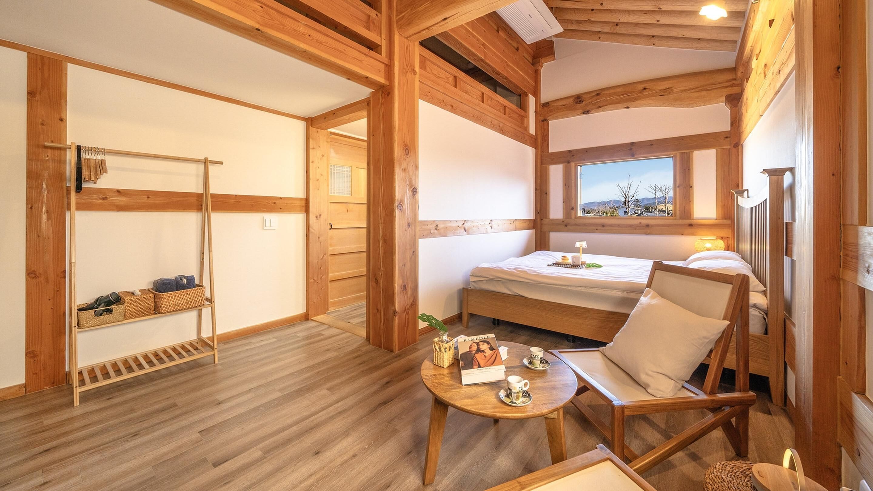 Ein Holzhäuschen im Inneren mit einem Bett, einem Sessel, einem kleinen Tisch und einem Fenster mit Blick.
