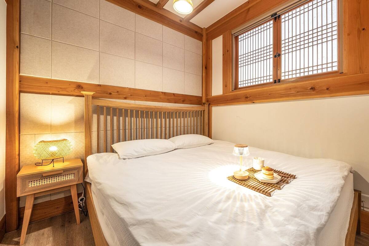 Ein Schlafzimmer mit hölzernem Kopfteil, einem Bett mit weißer Bettwäsche, einer Nachttischlampe und einem Fenster mit Holzrahmen.