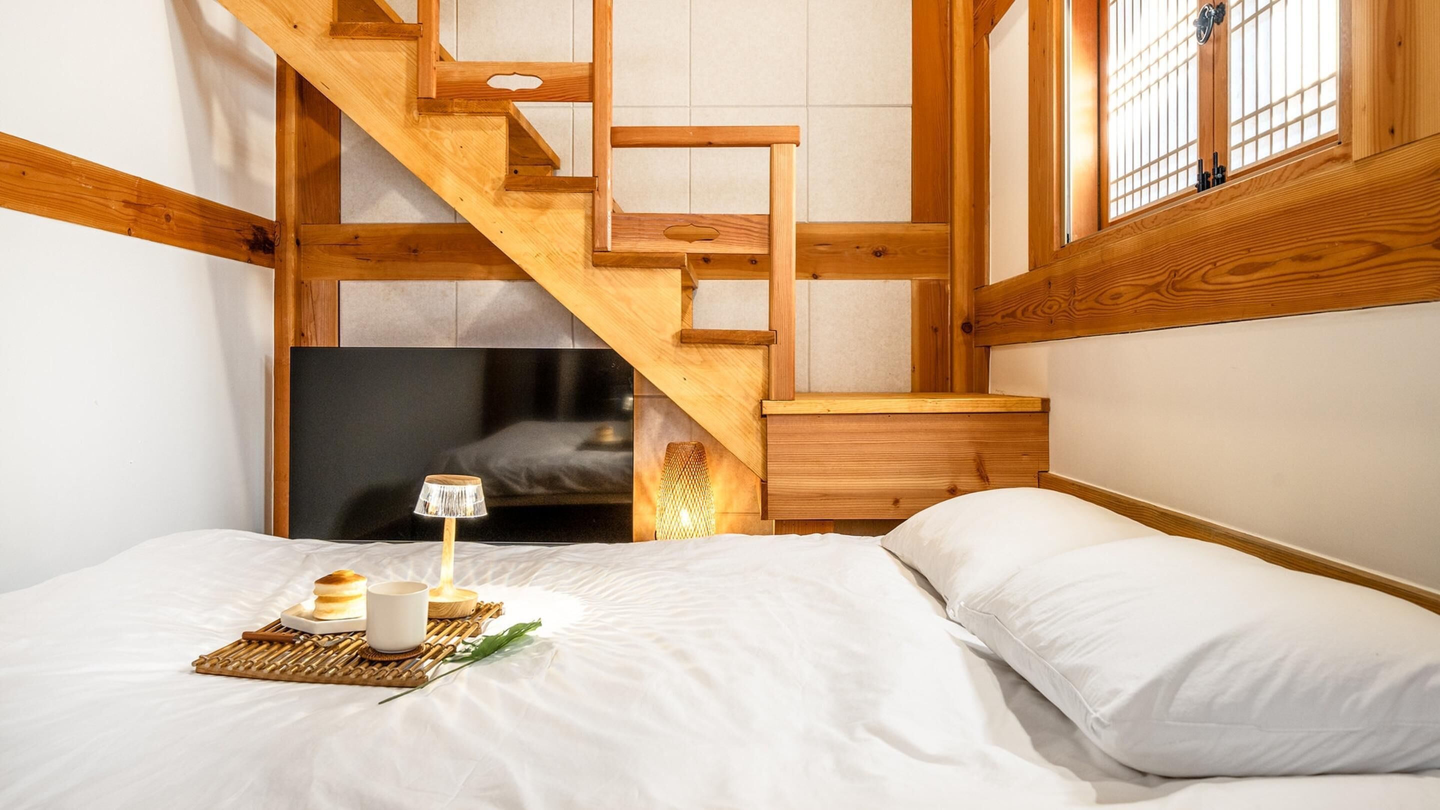 Un dormitorio con escalera de madera, cama con ropa de cama blanca, mesita de noche y ventana con barrotes.