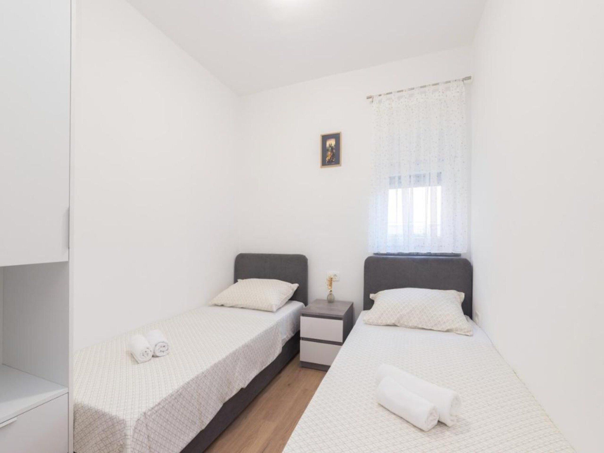 A small, clean room with two single beds, a nightstand, a small table, and a window with sheer curtains.