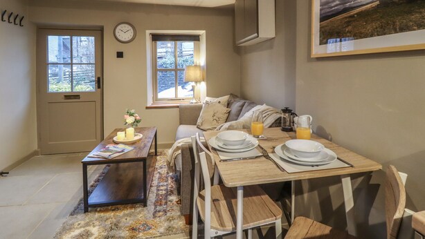 Une pièce chaleureuse avec une table à manger dressée pour deux personnes, un canapé, une horloge et une fenêtre donnant sur l’extérieur.