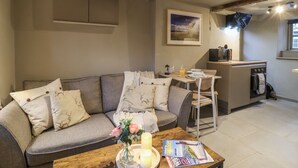 Un salon confortable avec un canapé, une table basse ornée d’un vase de fleurs et un magazine. Il y a également un coin cuisine équipé d’un four à micro-ondes et d’une fenêtre.