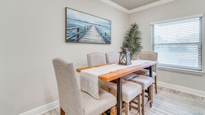 A dining area with a wooden table, beige upholstered chairs, and a painting of a pier on the wall. - Pensacola Vacation Rental w/ Community Pool! (Pensacola)