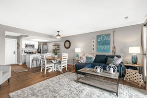 A modern living room with a blue sofa, a dining area with white chairs, a kitchen with white cabinets, and a large wall clock. - On Lake Time (Kimberling City)