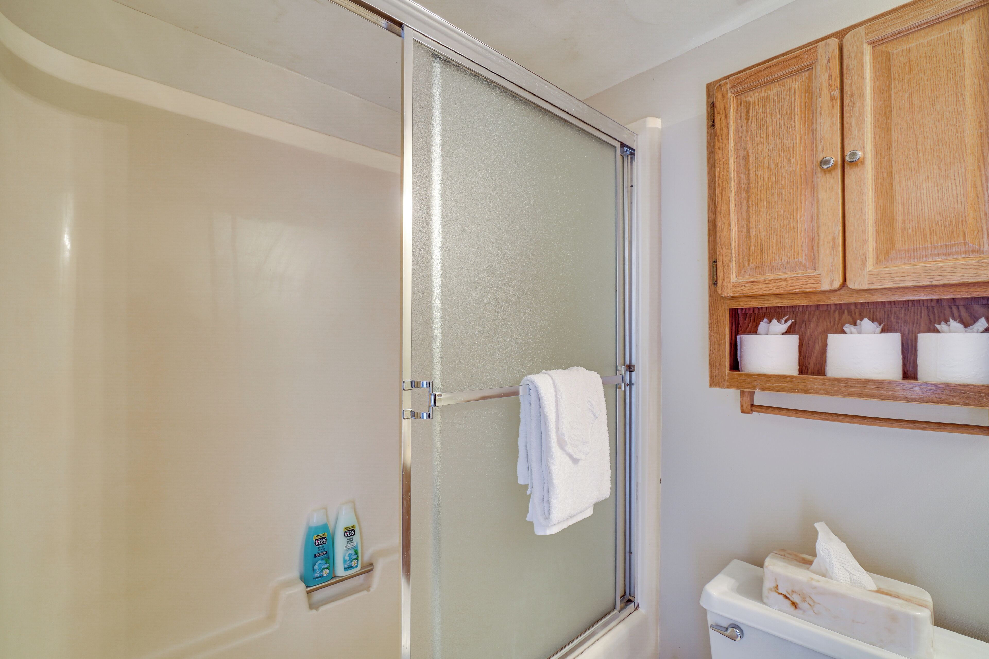 A bathroom with a glass shower enclosure, a white towel hanging on a rack, and a wooden cabinet above the sink with toiletries.