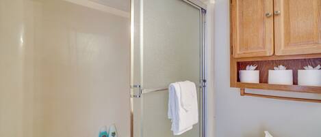 A bathroom with a glass shower enclosure, a white towel hanging on a rack, and a wooden cabinet above the sink with toiletries.
