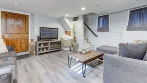 Un salon moderne avec un canapé gris, une table basse en bois, un téléviseur et un escalier adossé à un mur en briques blanches.