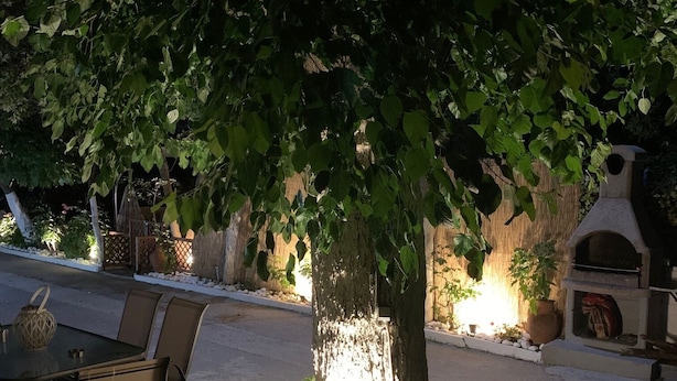 Zona esterna con tavolo e sedie all'ombra di un albero, circondata da un fioriera in pietra con rocce e un piccolo elemento d'acqua.