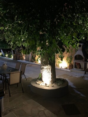 Un espacio al aire libre con mesa y sillas bajo un árbol, rodeado por una jardinera de piedra con rocas y un pequeño elemento acuático.