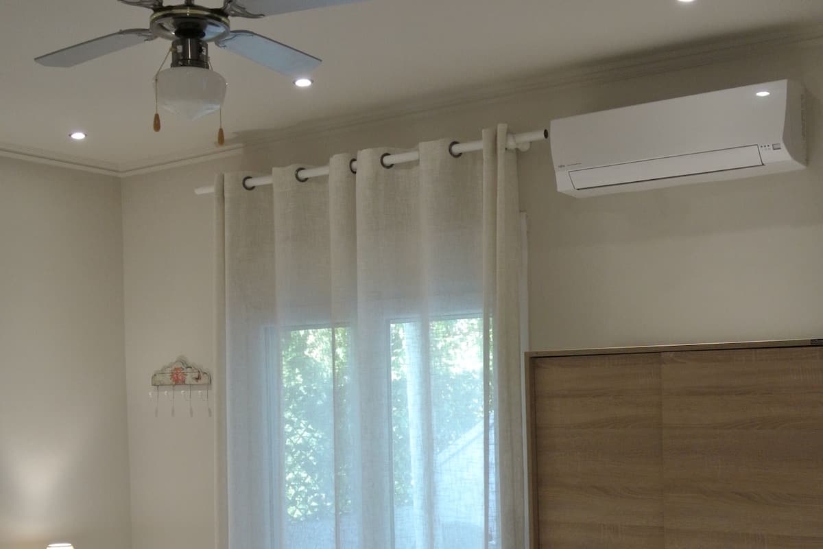 A bedroom with a bed, a ceiling fan, an air conditioning unit, a nightstand with a lamp, and a window with curtains.