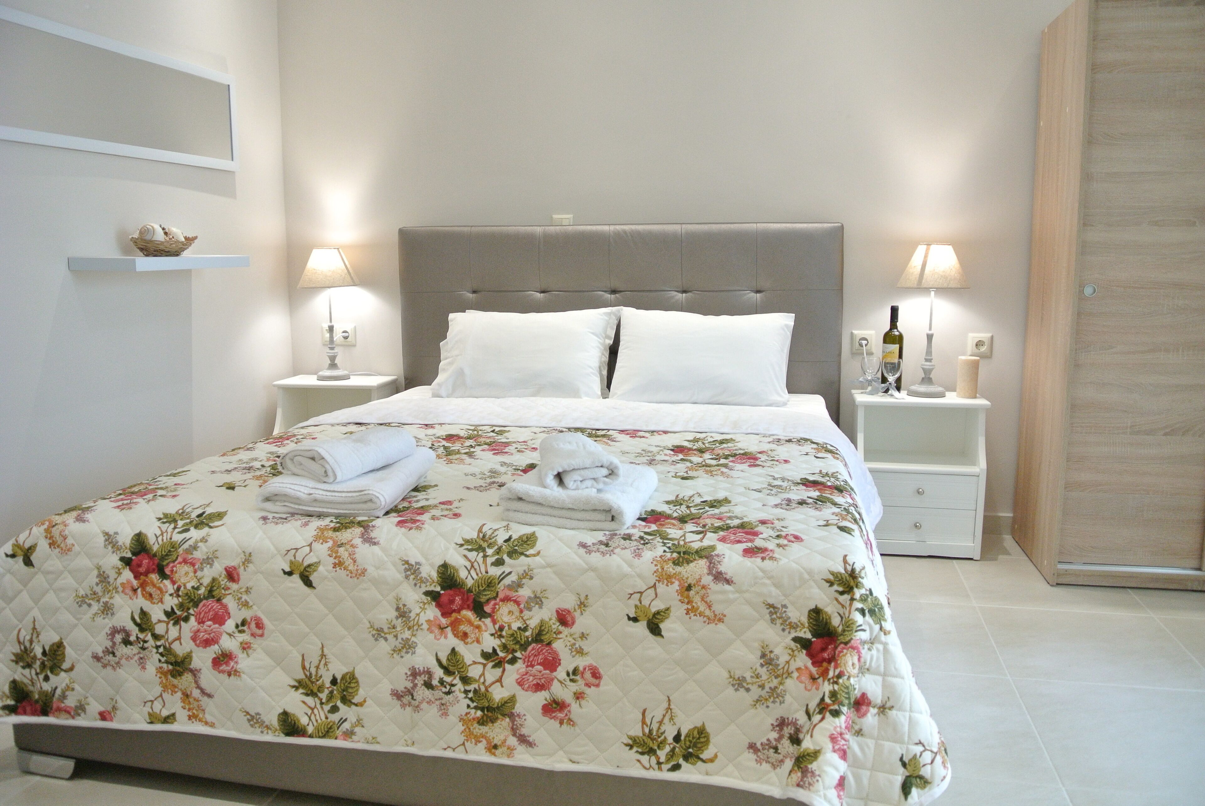 Un lit bien fait, recouvert d’une courtepointe à motifs floraux, deux tables de chevet avec des lampes et une petite étagère ornée d’objets décoratifs.