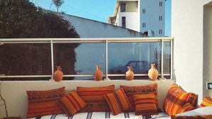 A sunlit room with a sofa set against a white wall, a window with vases, and a view of a modern building. - On the Beach you see Horses and Quad Riders (Essaouira)